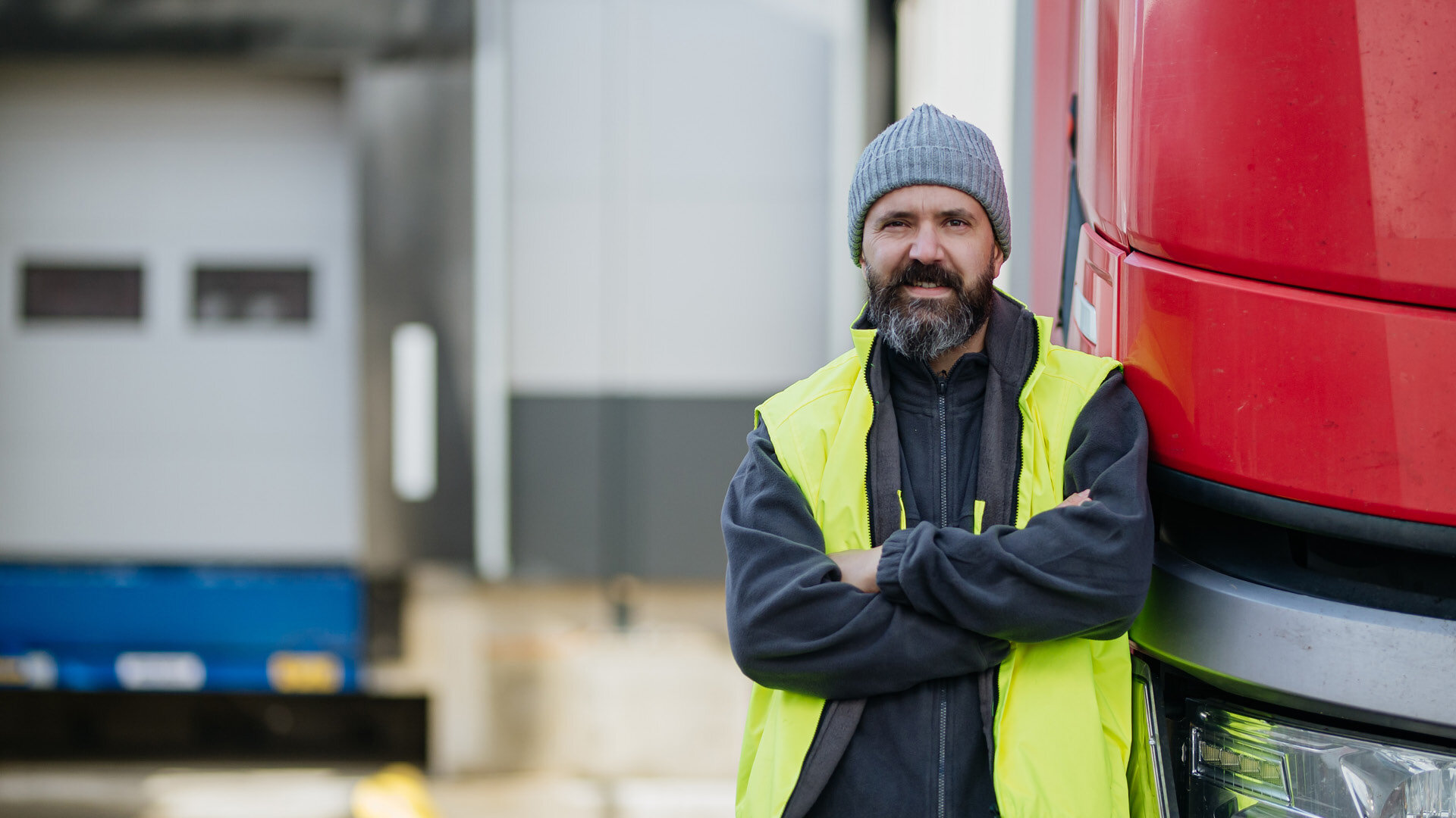 Mann mit Mütze und Warnweste lehnt an Lkw auf Parkplatz von Spedition